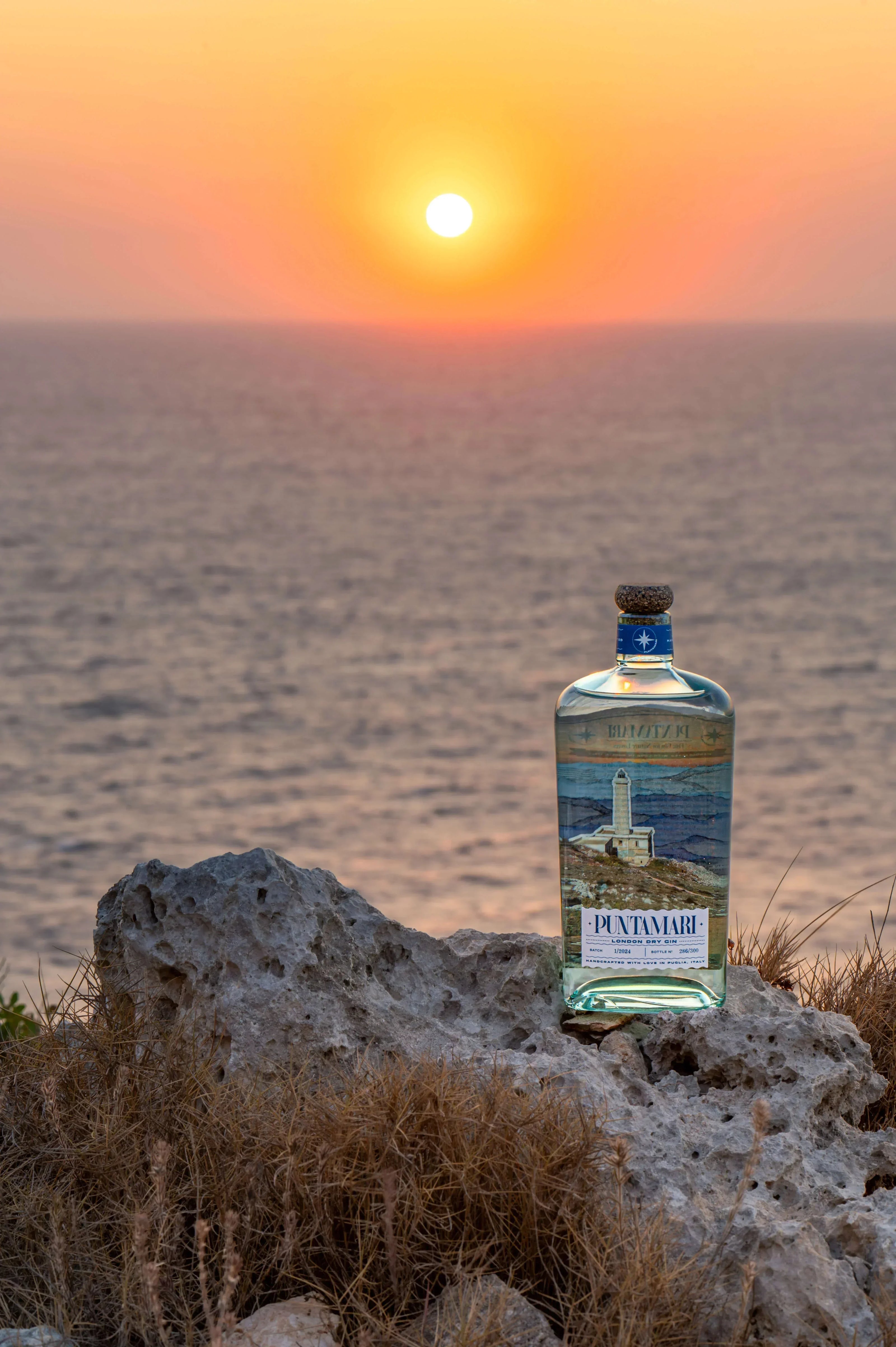 Bottiglia di Puntamari, gin artigianale italiano prodotto in Puglia, posizionata su una scogliera vista mare durante un tramonto mediterraneo